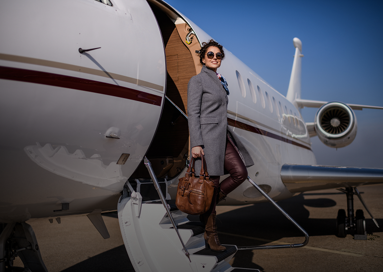 Woman standing outside private jet, holding bag.
