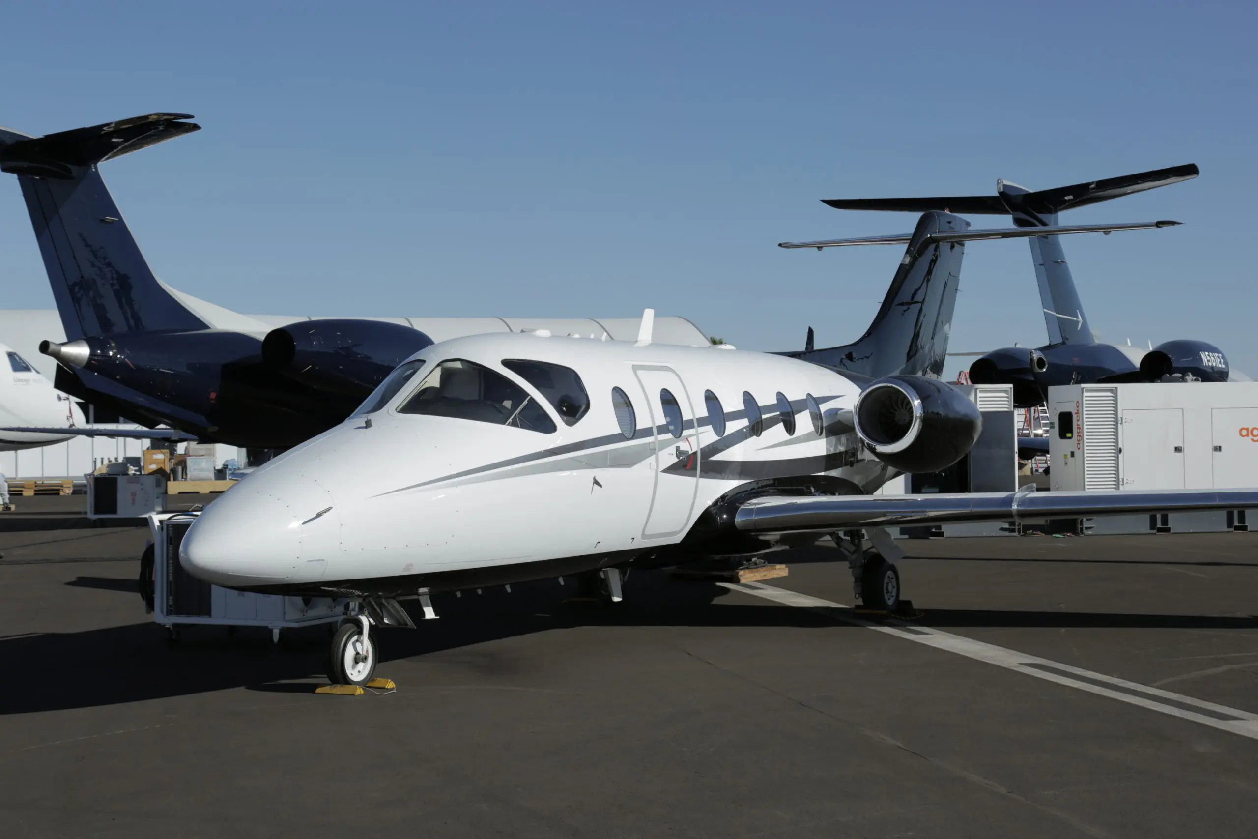 Small white private jet parked on tarmac with larger planes in background.