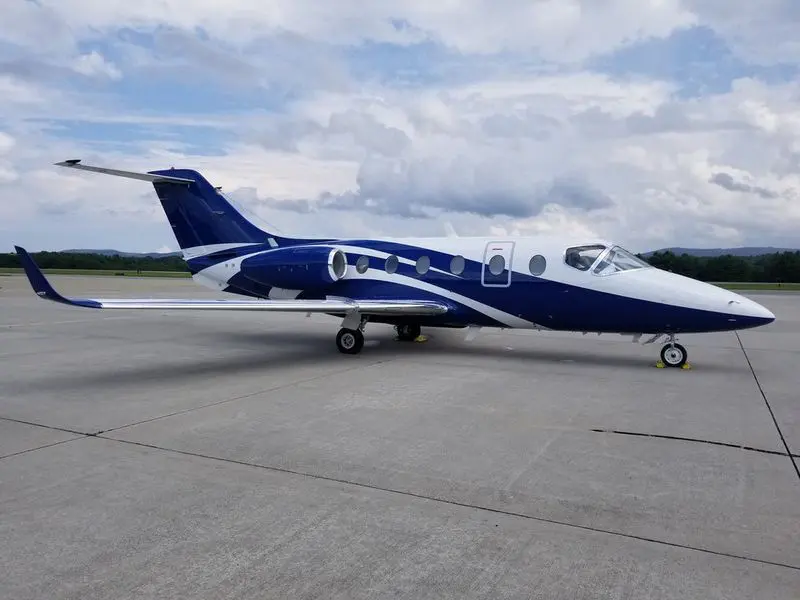 A small blue and white private jet on an airport tarmac under a cloudy sky.