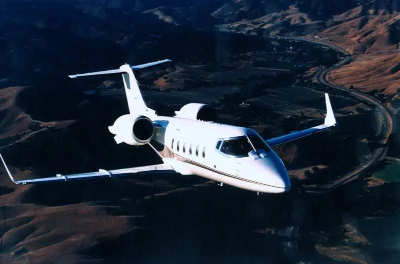 White private jet flying over mountainous terrain during sunset.
