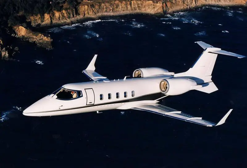 White private jet flying over water near cliffs.