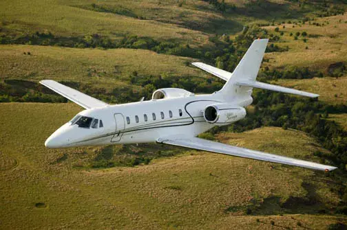 A sleek white private jet flying over green fields.