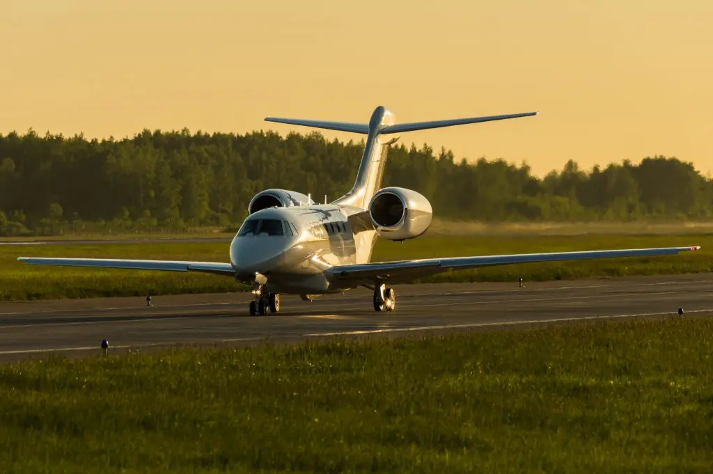 Private jet taxiing on runway at sunset with forest backdrop.