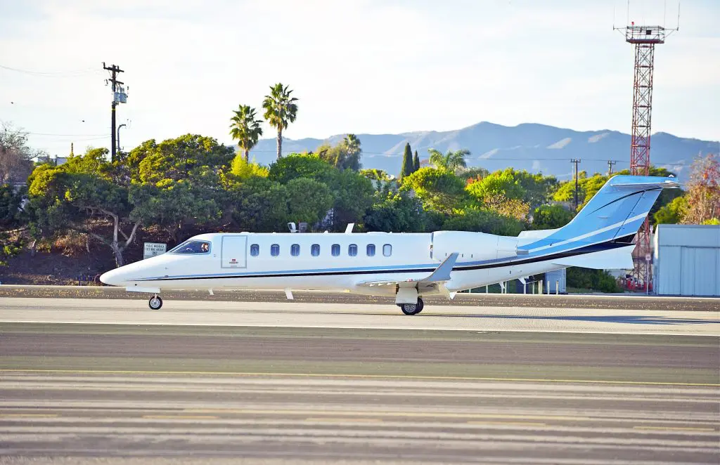 Private jet taxiing on runway with palm trees and mountains in background.