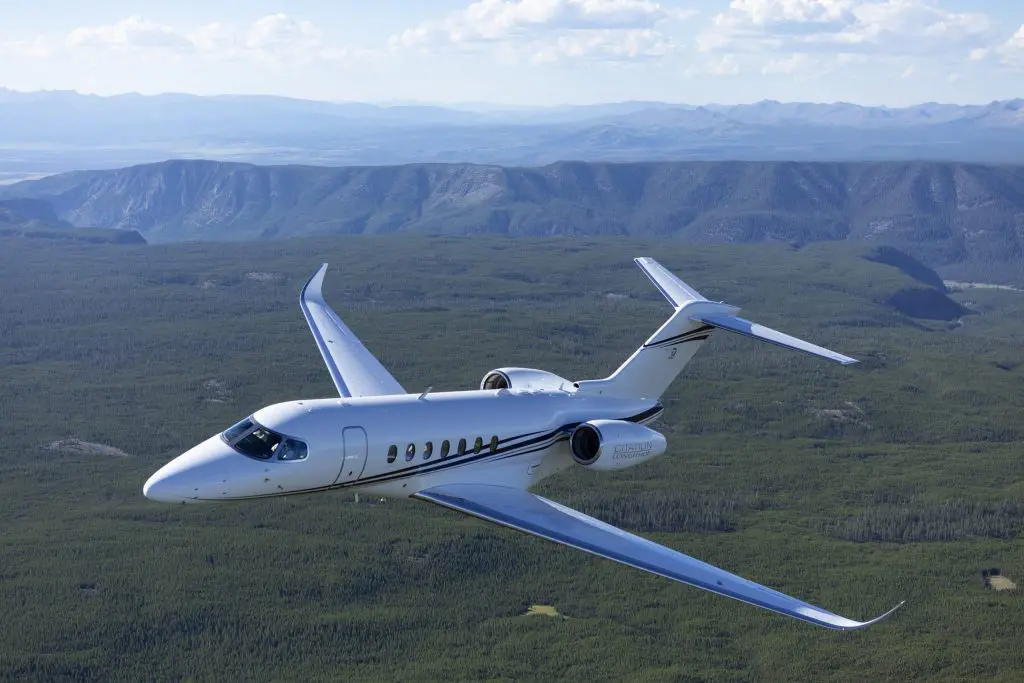 A sleek private jet flying over a vast green landscape with mountains in the distance.