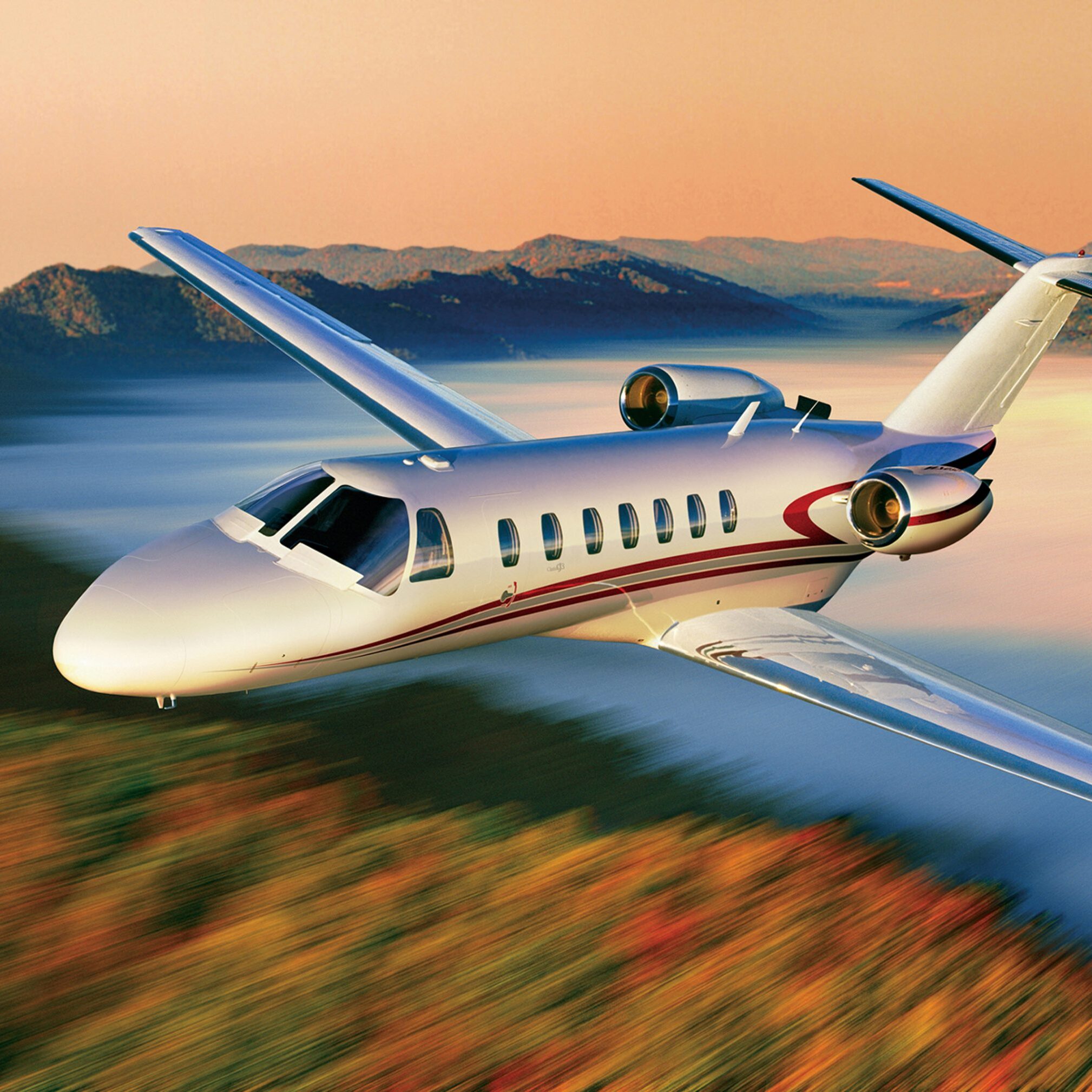 A sleek private jet flying over a mountainous landscape at sunset.