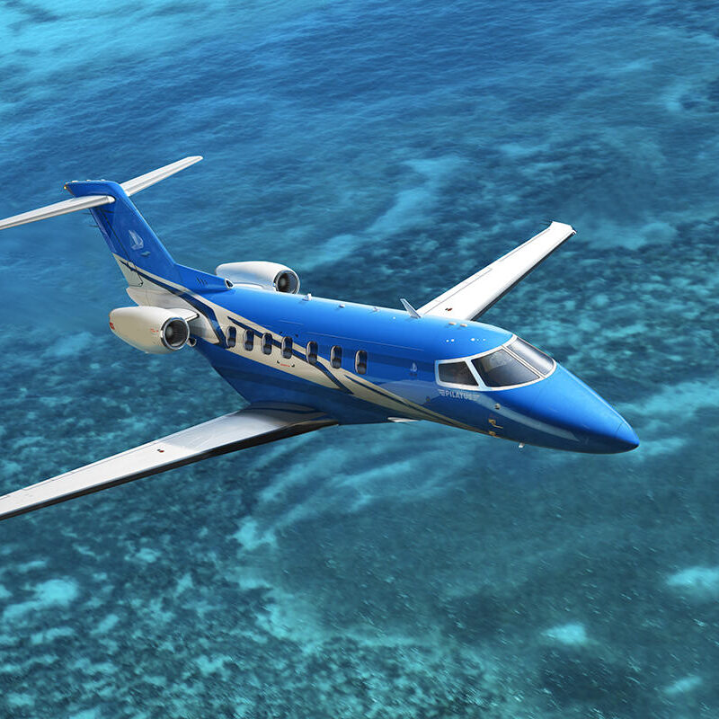 A sleek blue private jet flying over clear turquoise waters.